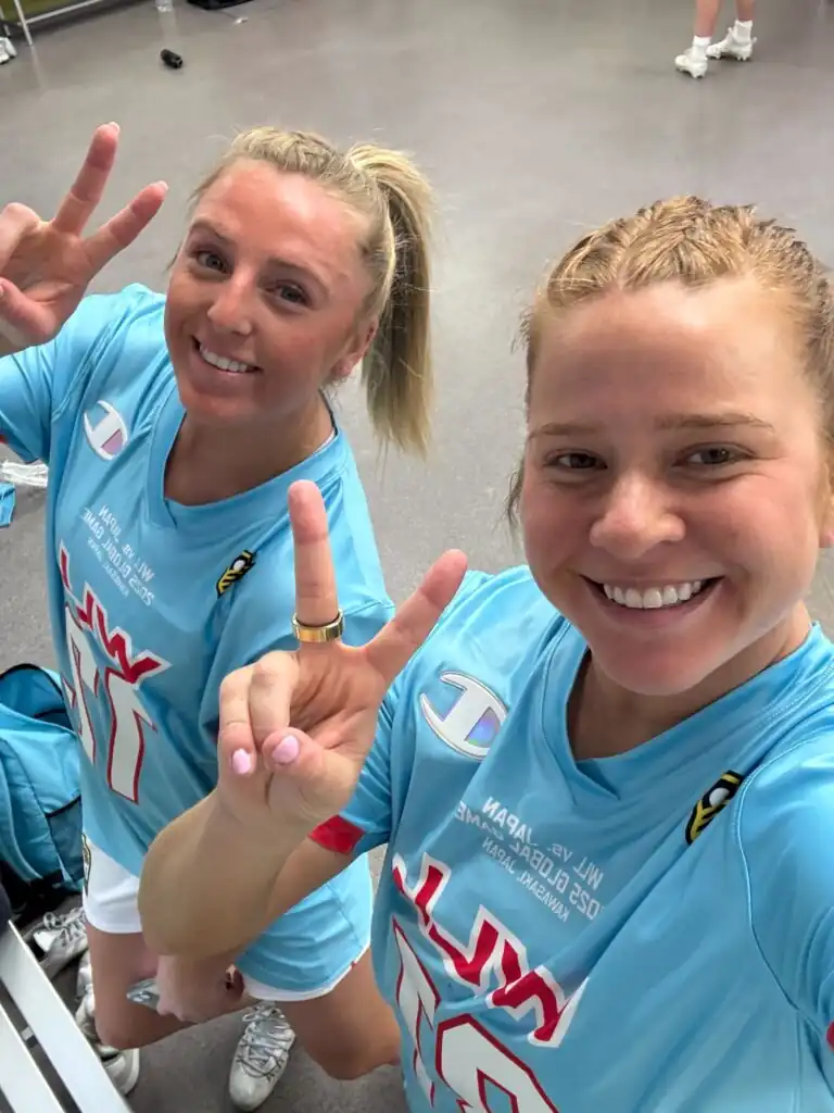 Two women in matching blue sports jerseys pose for a selfie, making peace signs with their fingers and smiling.
