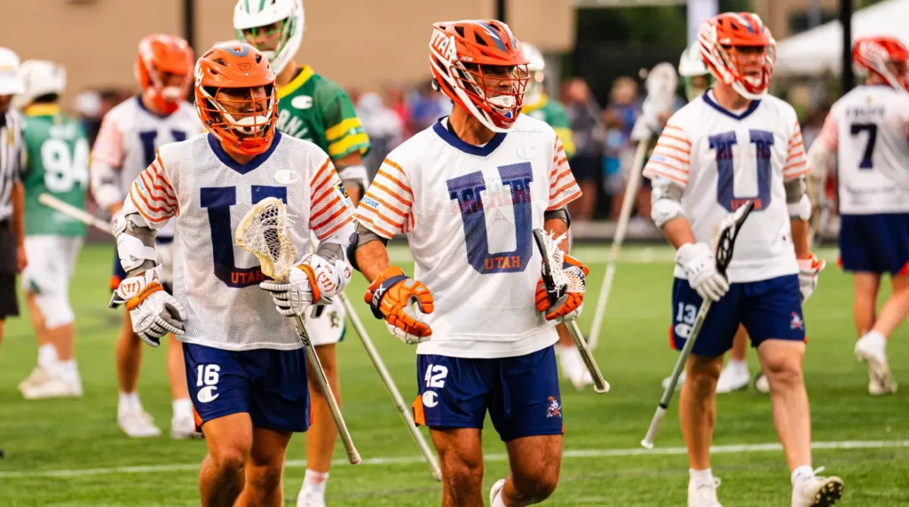 Lacrosse players in white and blue uniforms with orange helmets walk on the field, preparing for a game.