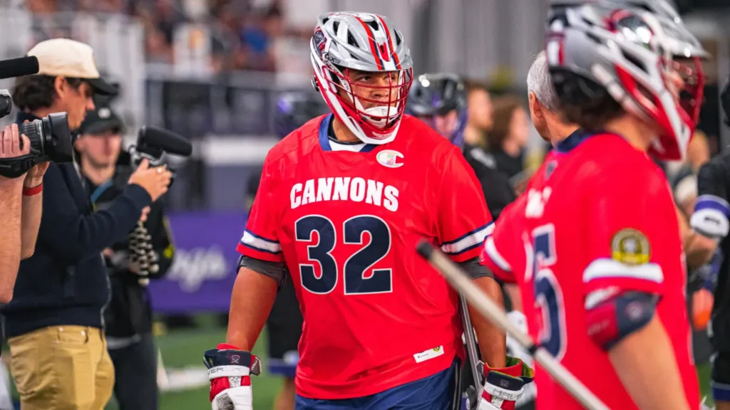 A lacrosse player in a red jersey with "CANNONS" and the number 32 walks on the field, surrounded by teammates and photographers.