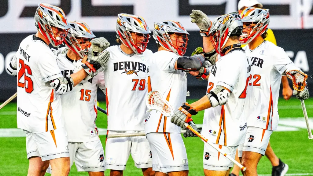 A group of lacrosse players in white jerseys celebrate together on the field, showcasing teamwork and excitement.