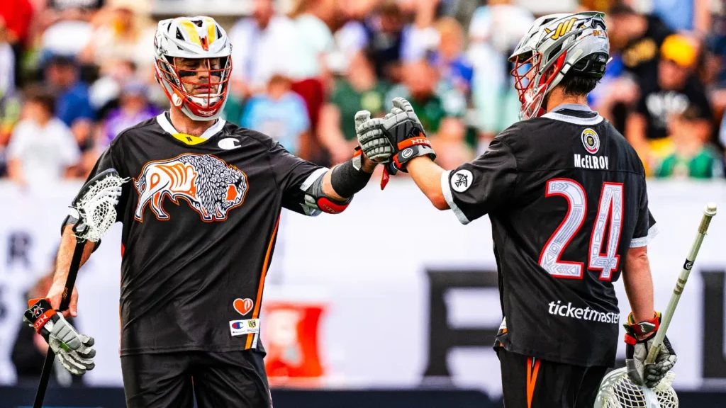 Two lacrosse players in black jerseys celebrate with a fist bump on the field, surrounded by a cheering crowd.