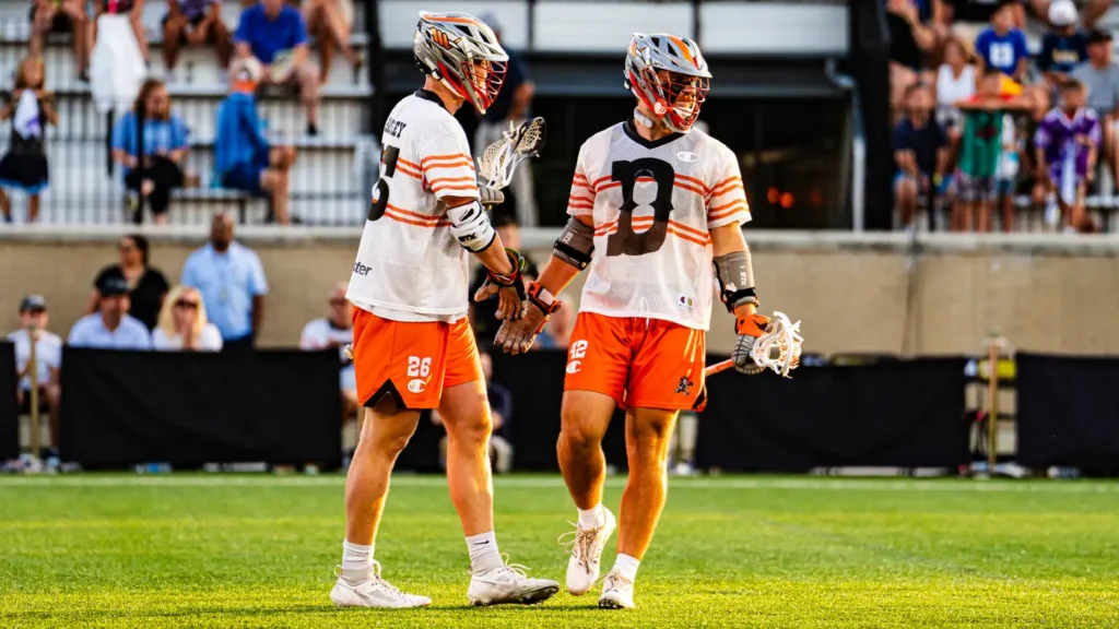 Two lacrosse players in orange and white uniforms are standing on the field, engaged in conversation. Spectators are visible in the background.