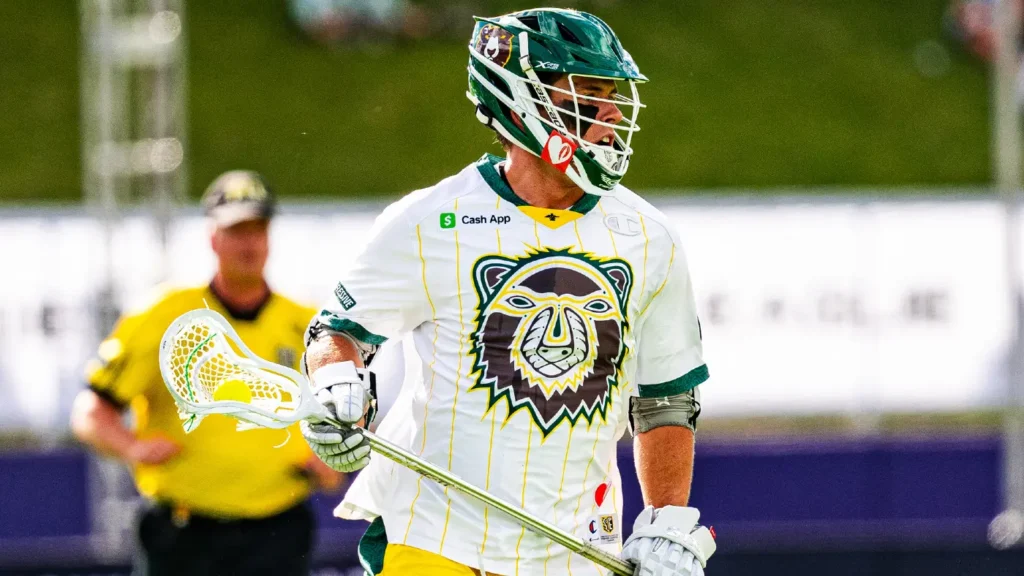 A lacrosse player in a white jersey with a bear logo, holding a lacrosse stick, during a game.