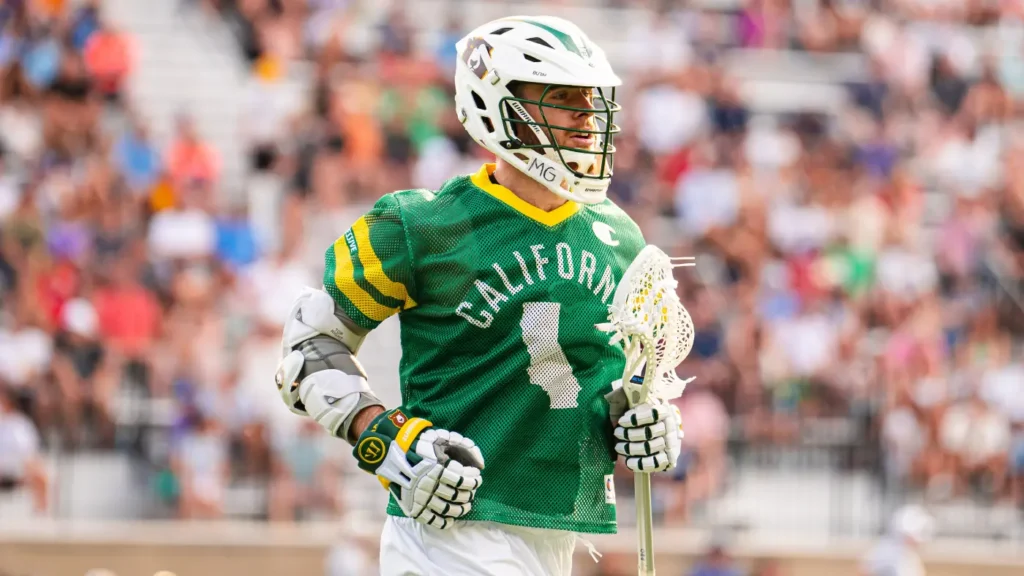 A lacrosse player in a green jersey with "CALIFORNIA" and the number 4, holding a lacrosse stick, stands on a field with a crowd in the background.