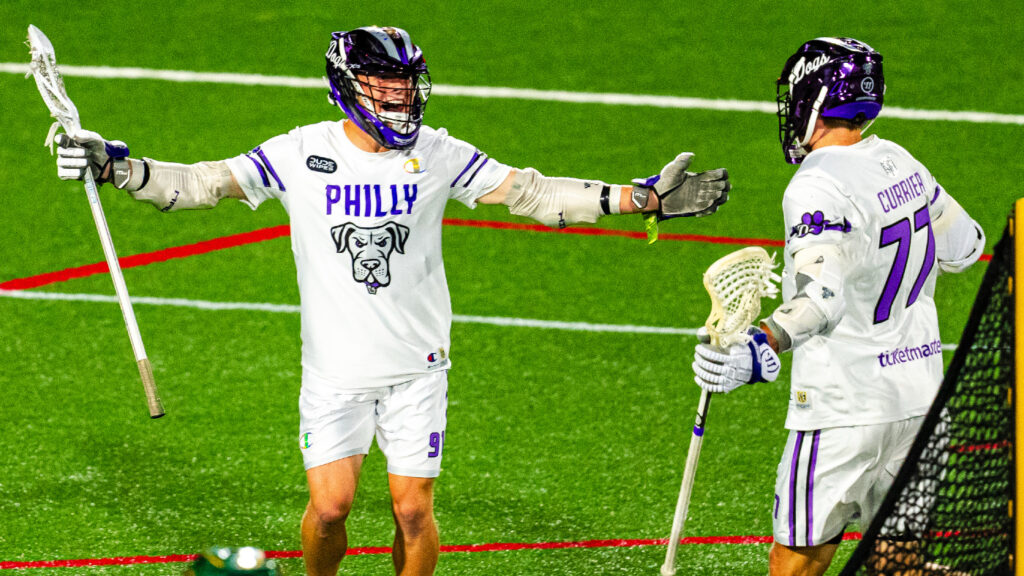 Two lacrosse players celebrate a goal on the field, wearing white jerseys with "Philly" and a dog logo.