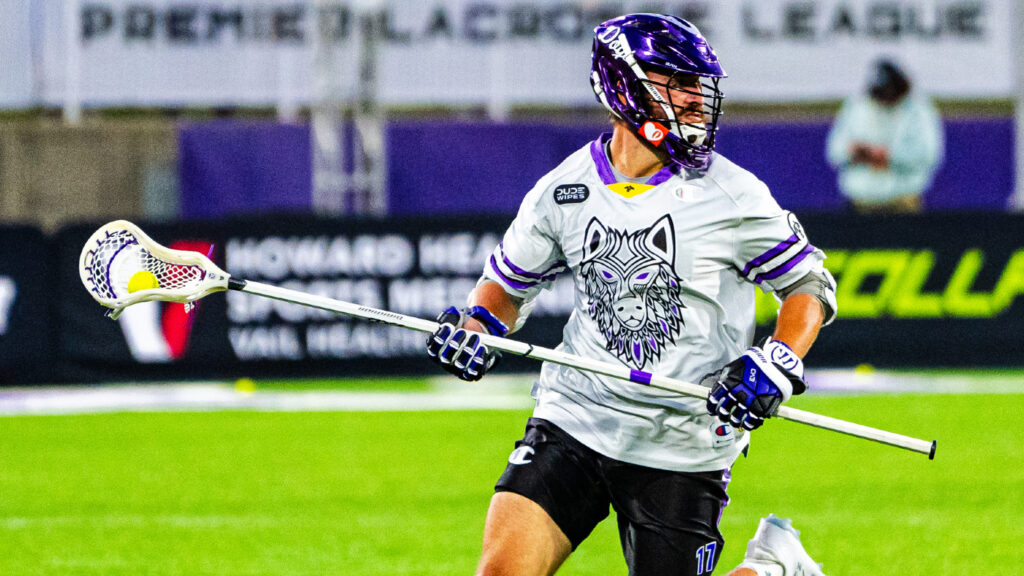 A lacrosse player in a gray jersey with a wolf graphic runs on the field, holding a stick with a ball in the net.