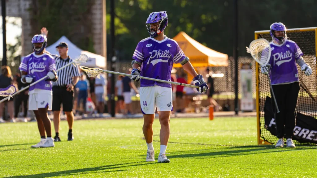 Lacrosse players in purple jerseys on a field, with a referee and spectators in the background.