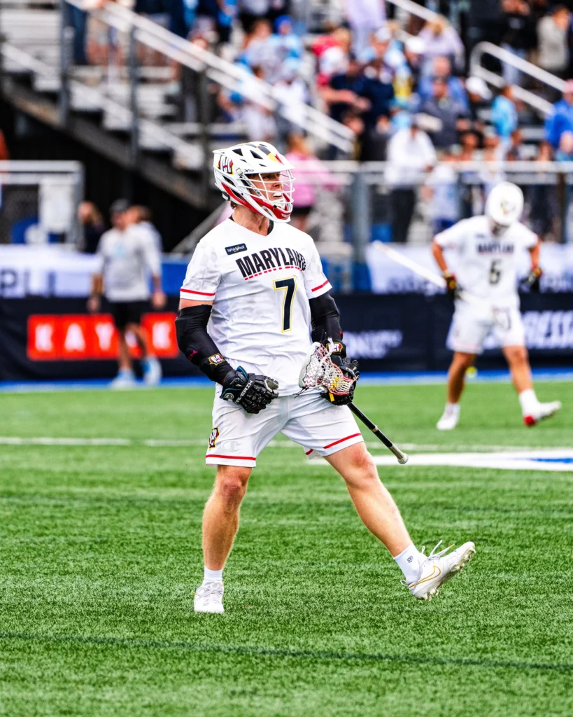 A lacrosse player in a white Maryland jersey with the number 7 stands on a field, holding a stick, with spectators in the background.